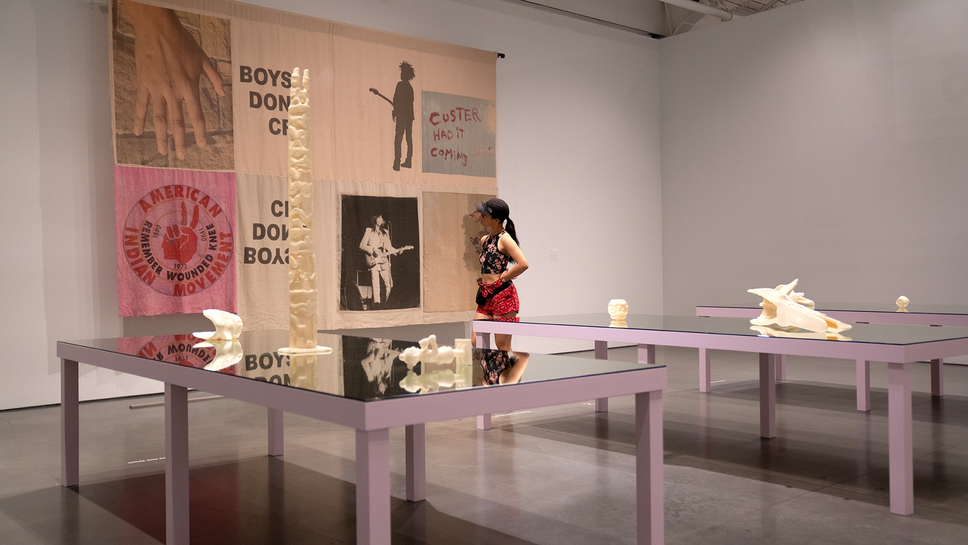 in background: a person in an gallery looks closely at a large linen artwork hanging on the wall, which has the words "boys don't cry" and "custer had it coming"; in foreground: eight cream-colored 3D printed objects sit on dark mirrored tables