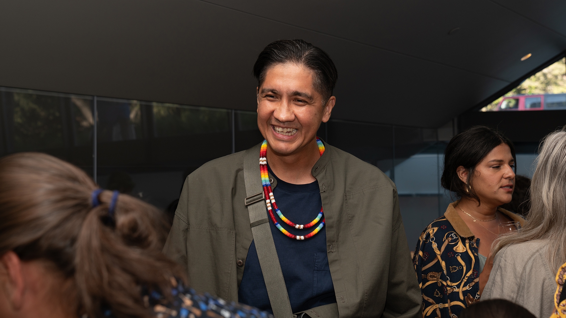 a person with short dark hair wearing a bright beaded necklace smiles at a person out of the frame