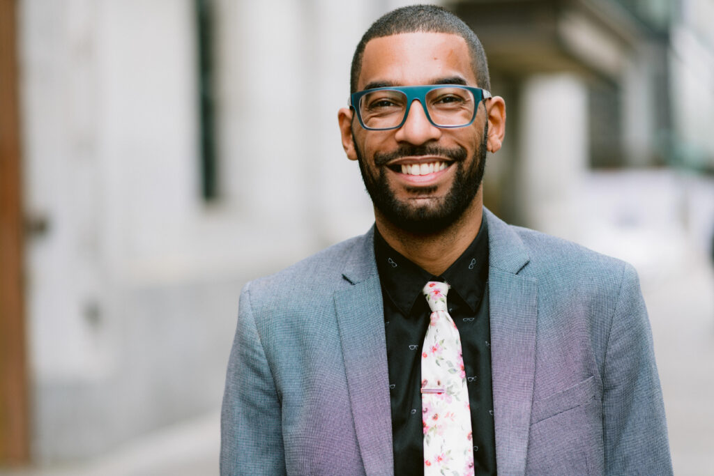 Christopher Williams wearing a grey suit and green eye glasses smiling.