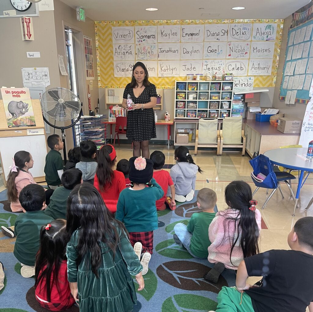 Leslie Cortes Sanchez stands in a classroom teaching her students as they sit in front of her attentively listening with the back of their heads to the camera.