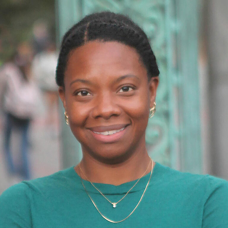 Tolani Britton wearing a green shirt with gold necklace and earrings smiling in front of a blurred Sather Gate in the foreground.