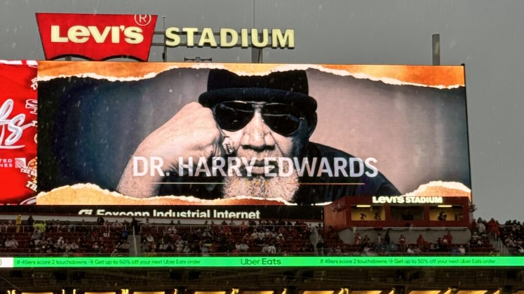 An image of Harry Edwards resting his right cheek on his fist is projected on a huge jumbotron screen at a 49ers football game.