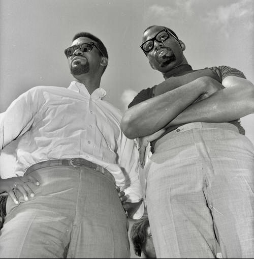 Black and white photo of Harry Edwards standing with arms folded, next to Malcolm X.