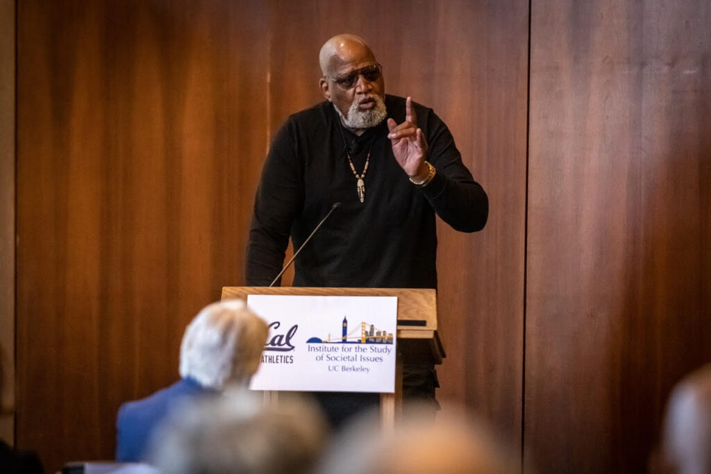 Harry Edwards speaking behind a podium and gesturing with his hand to a group during an event at UC Berkeley in 2022