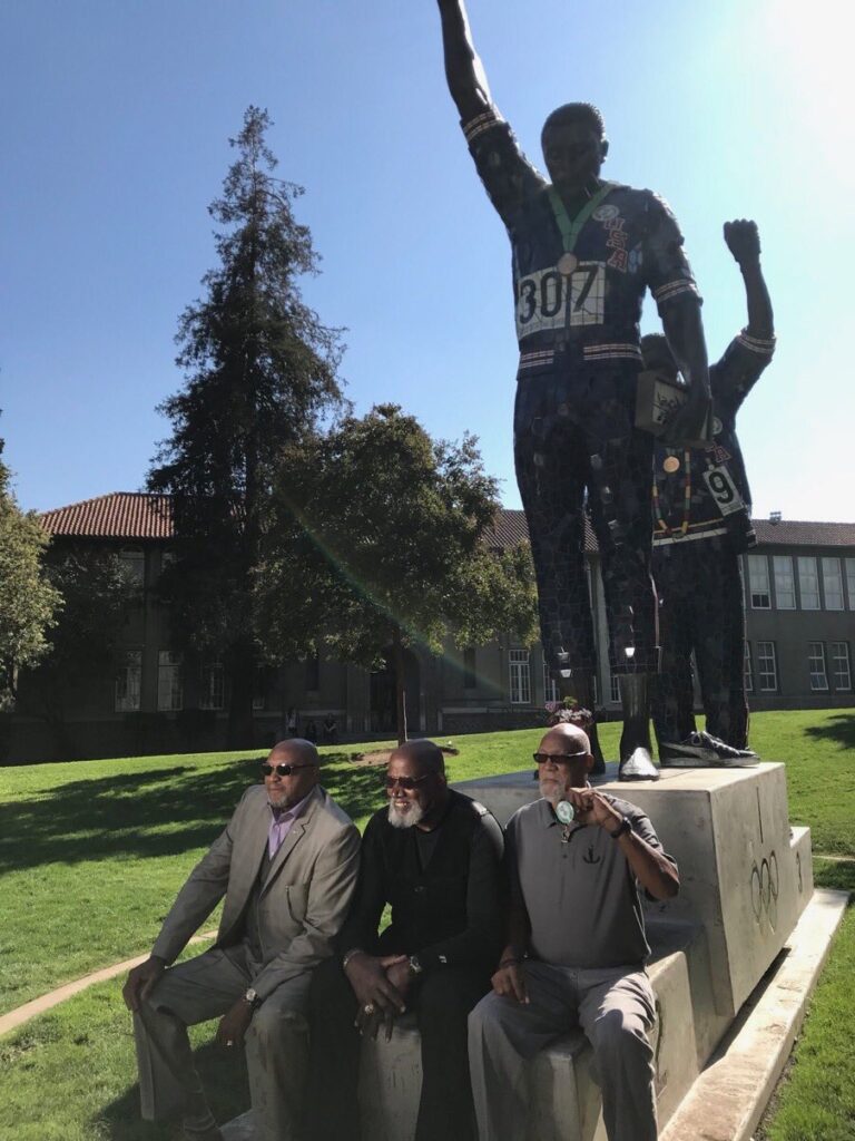 Harry Edwards sits on a paved stoop with Tommie Smith and John Carlos  just below a statue of two Olympians raising their fists while standing on a podium.