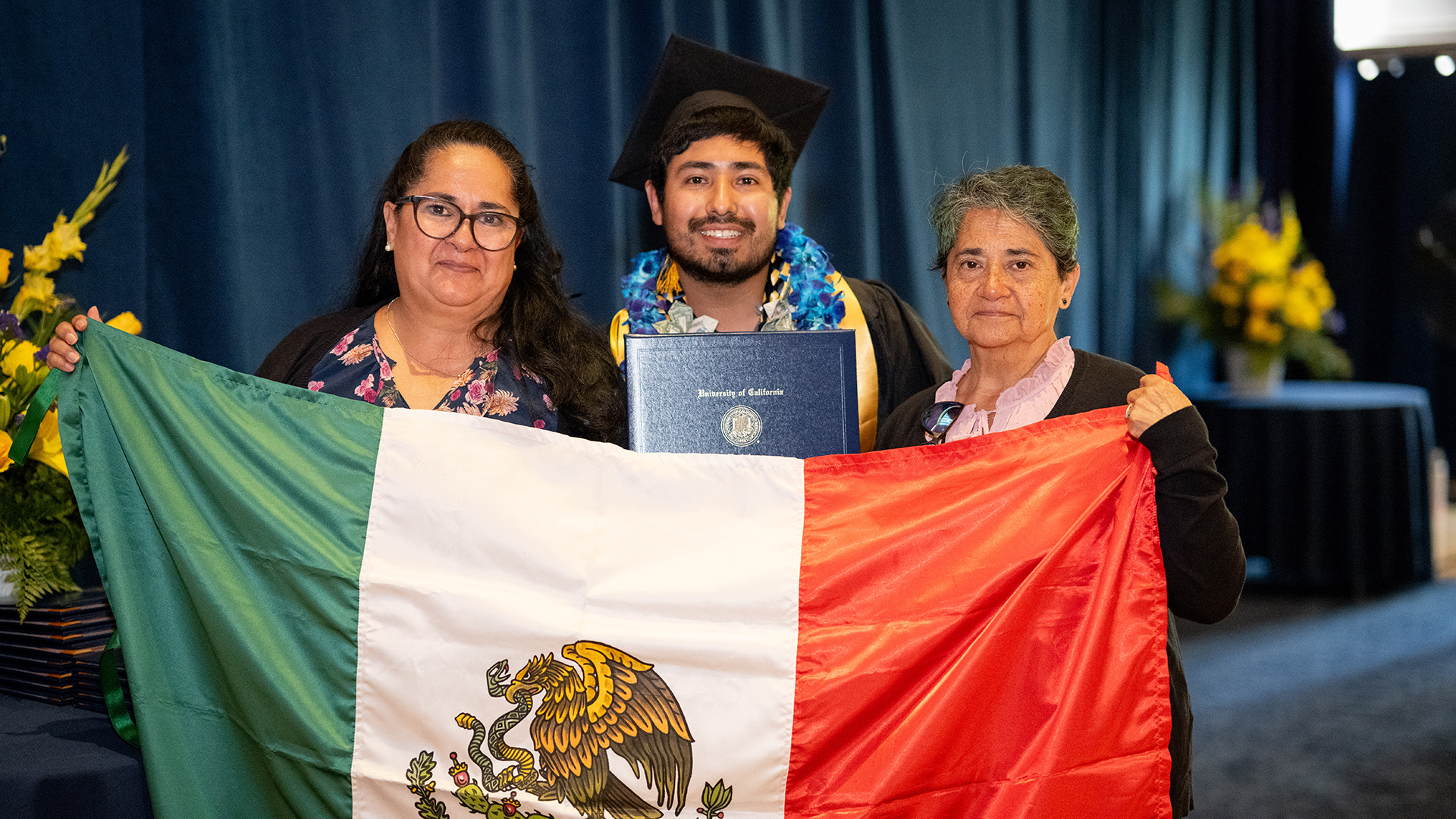 A student wearing a graduation cap and gown with two family members on either side smile and hold up a Mexican flag
