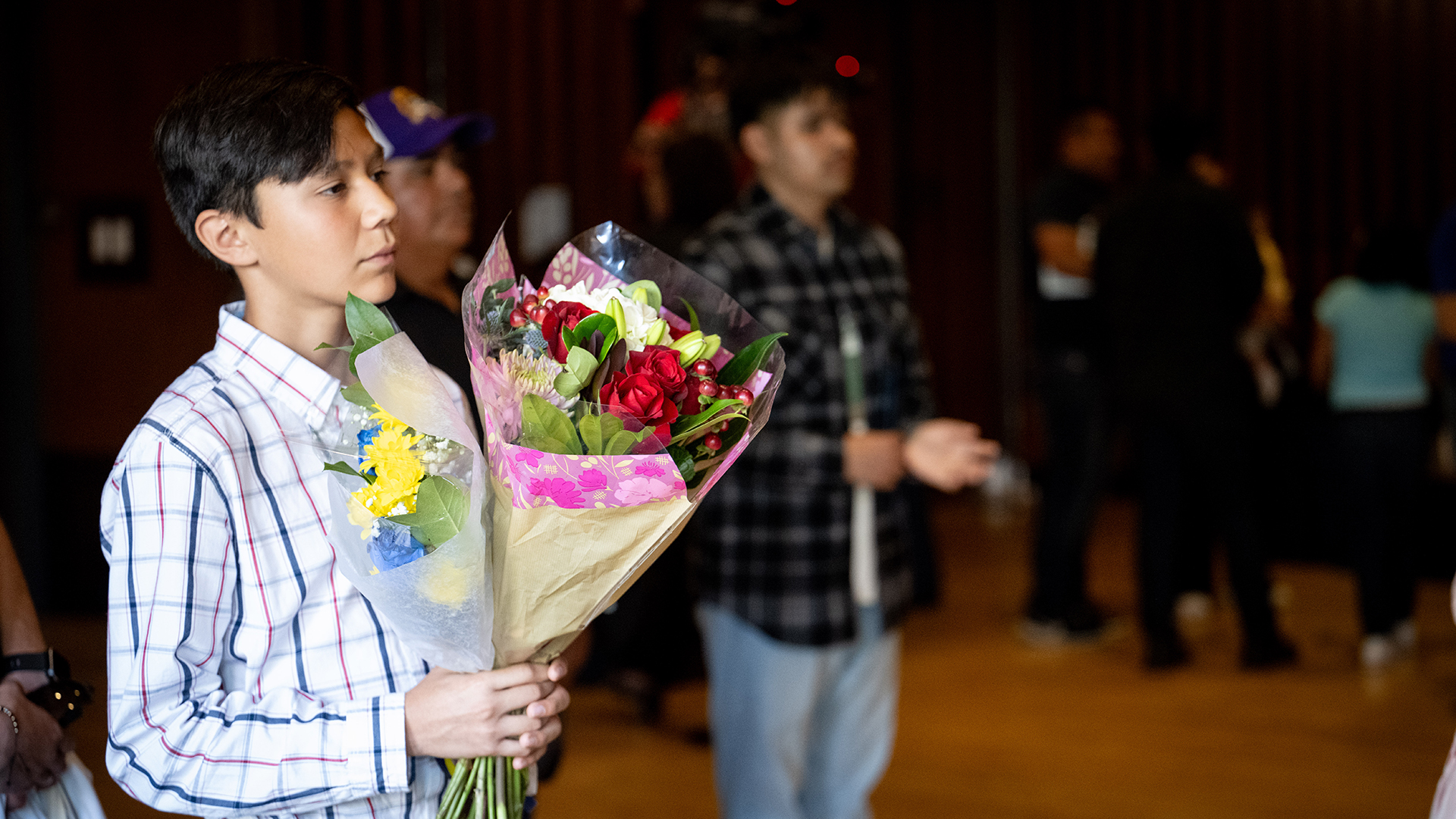 A teenager holds two bouquets of flowers and looks toward someone out of the frame during a graduation ceremony
