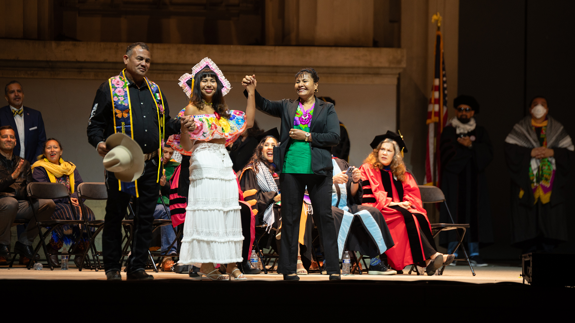 A graduating student stands on stage smiling and holding hands with her parents