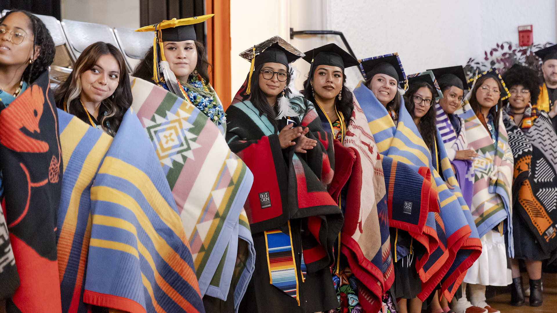 Graduating Native students stand in a line wearing their graduation caps and gowns and gorgeous Native blankets wrapped around their shoulders