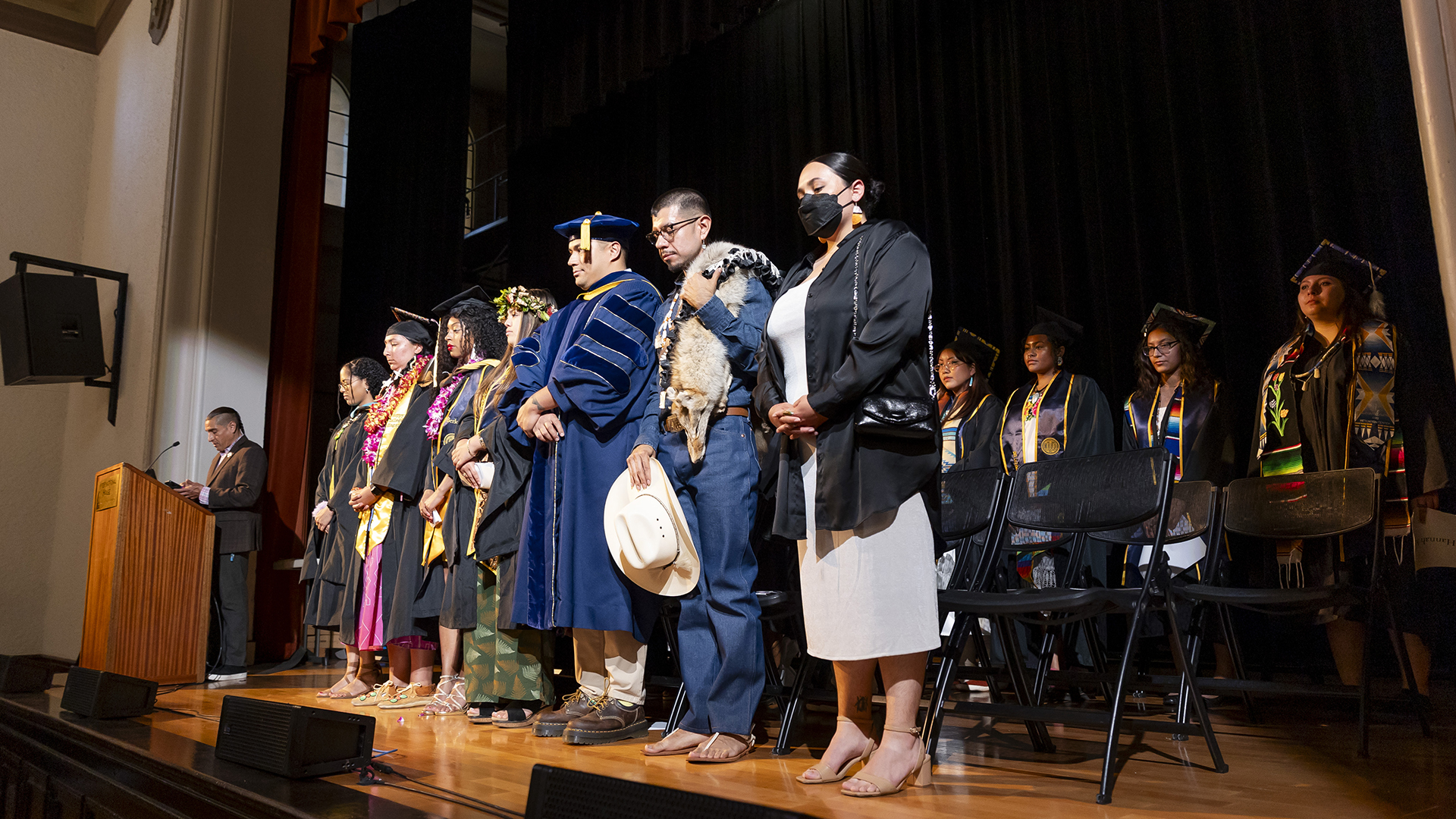 A little over a dozen Native graduates and others stand on stage during a graduation ceremony