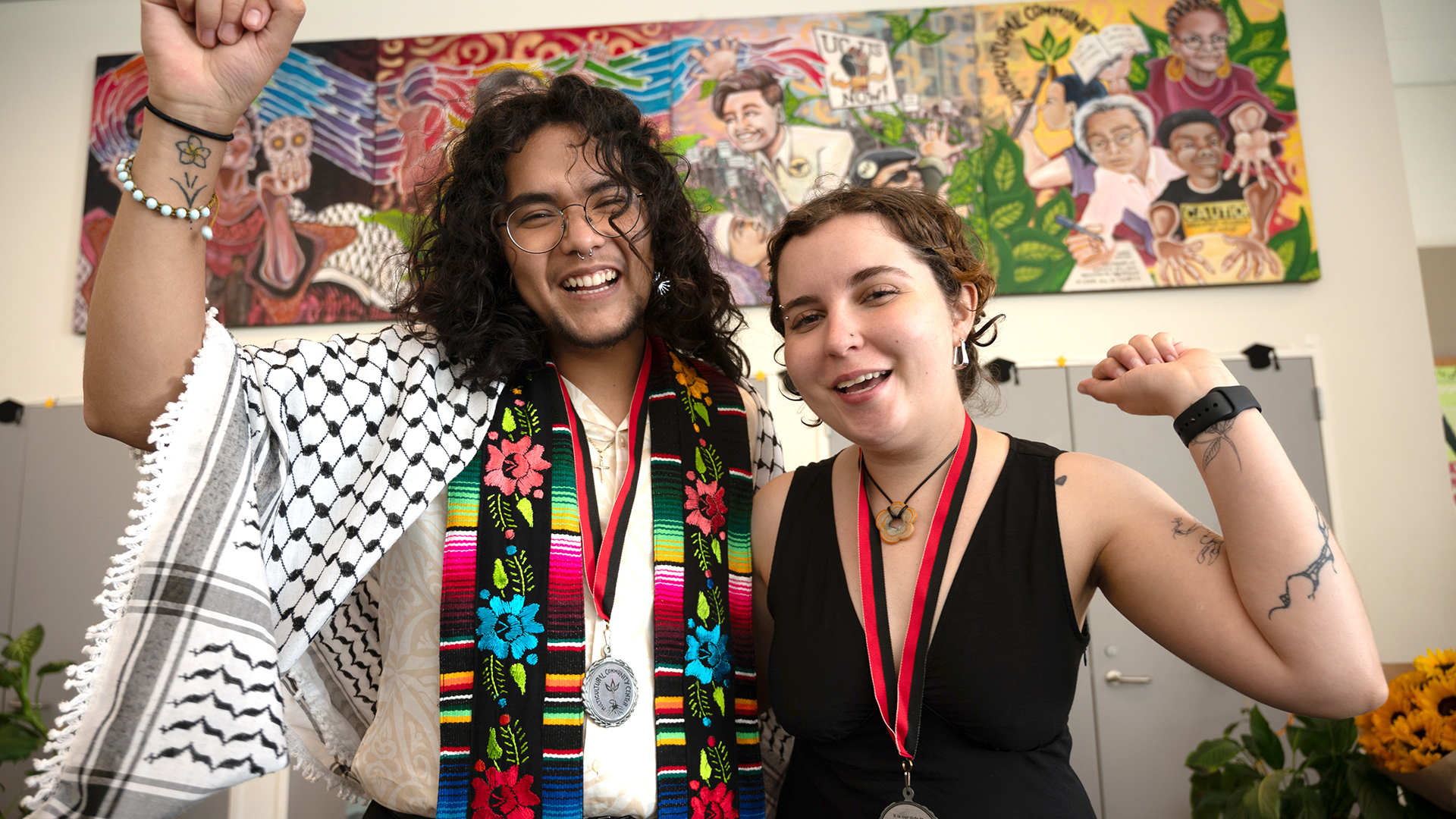 Two graduating students standing close each hold up a celebratory fist in the air and smile