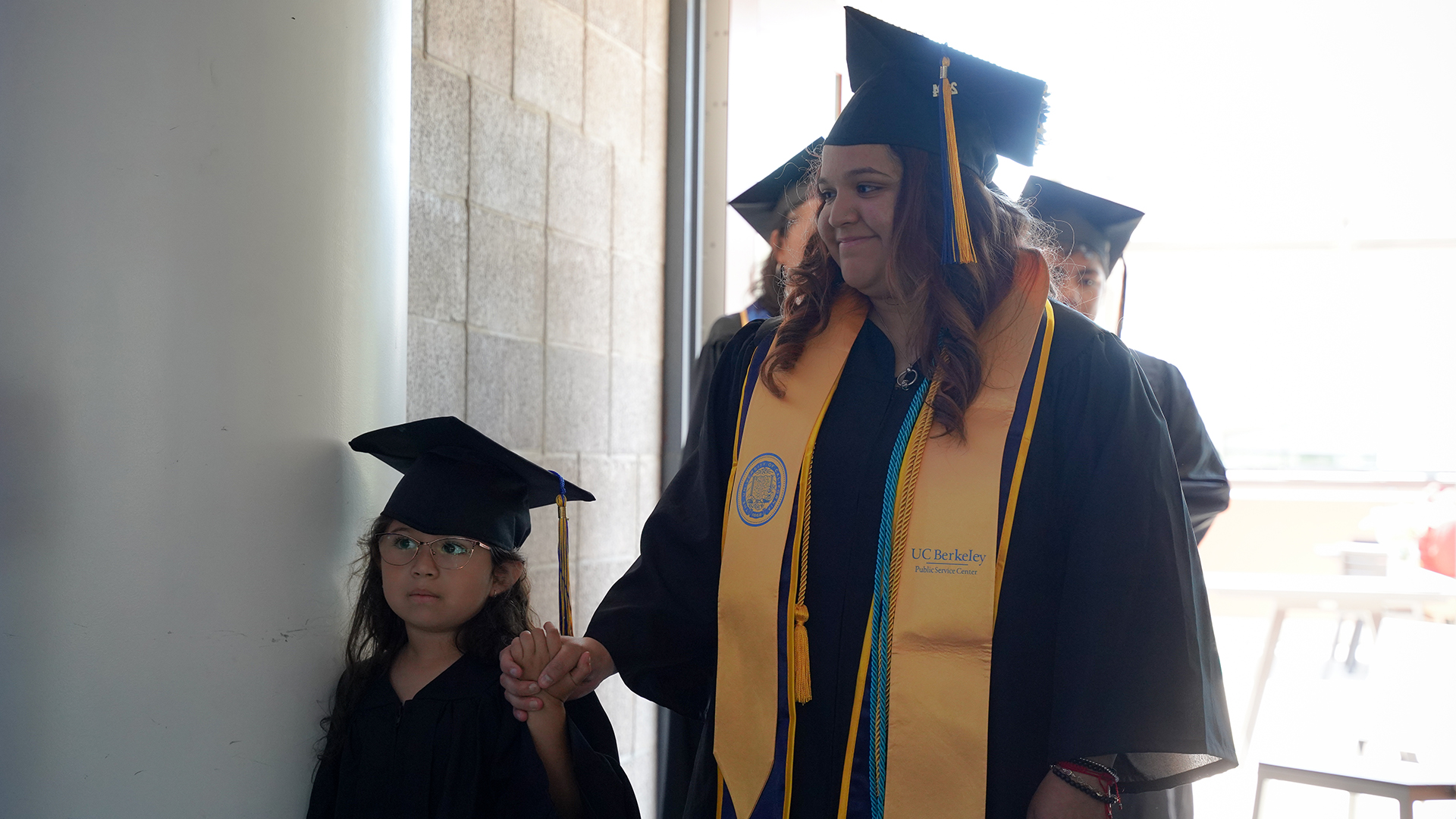 A graduating student in a cap and gown holds her daughter's hand, also wearing a little cap and gown