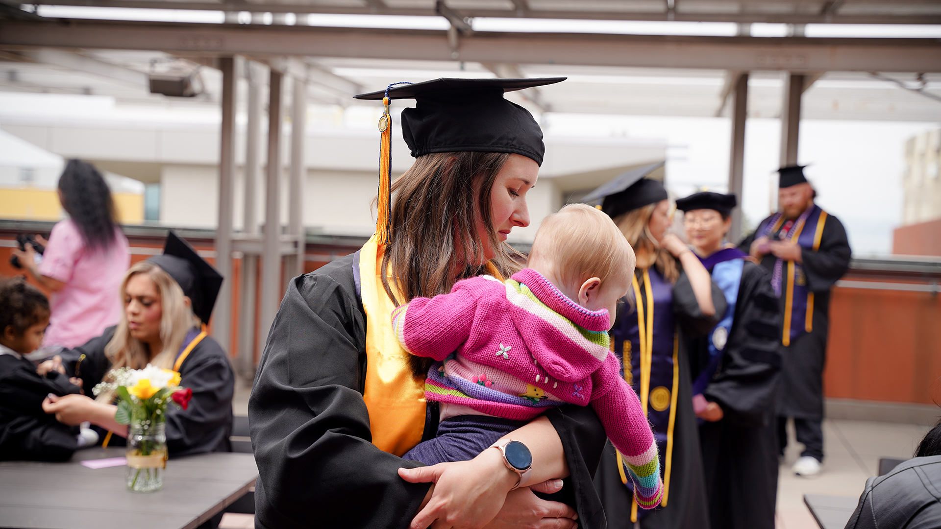 A graduating parent wearing a cap and gown holds her baby