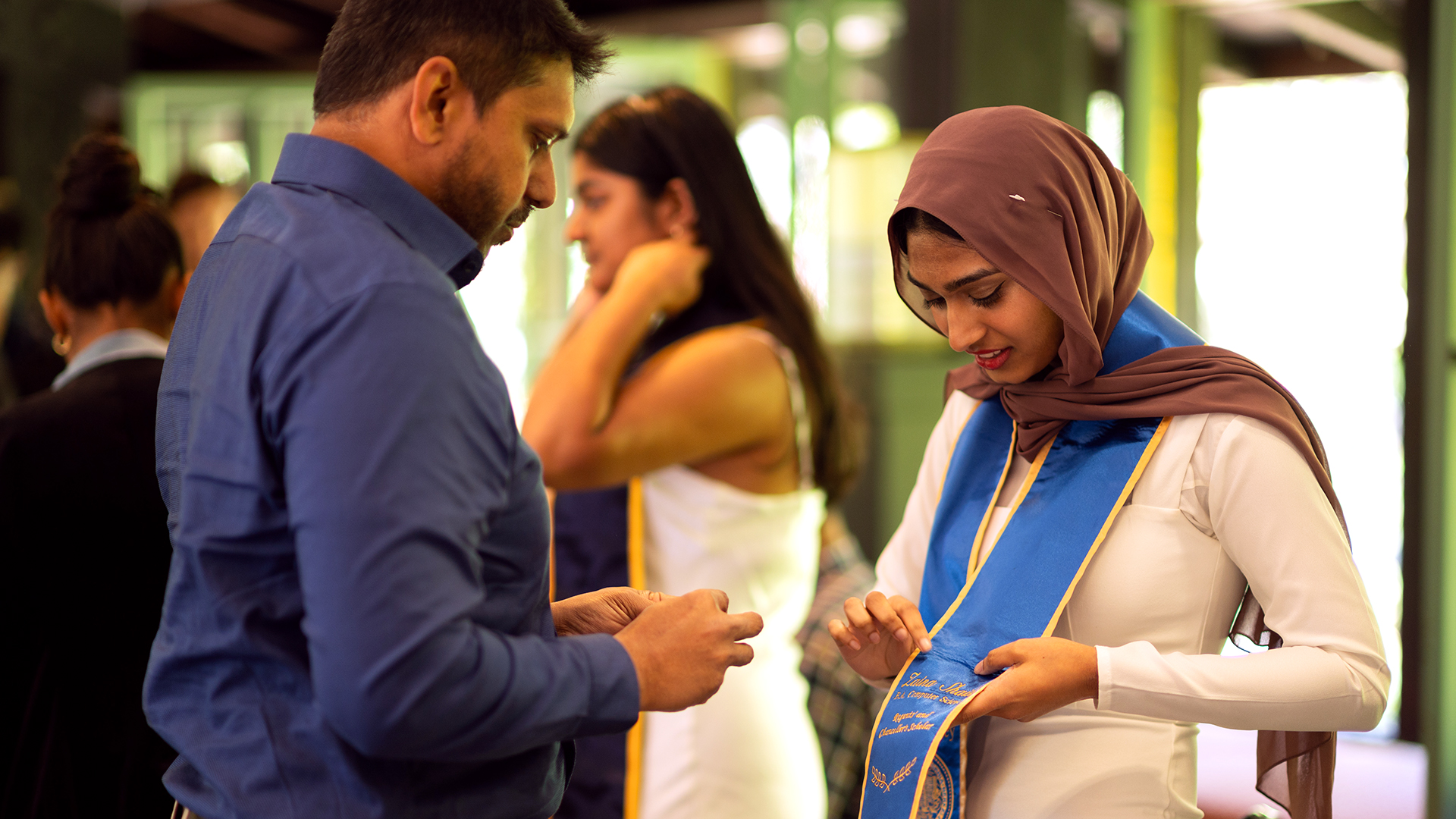 A graduating student wearing a headscarf holds out her graduation sash for it to be pinned