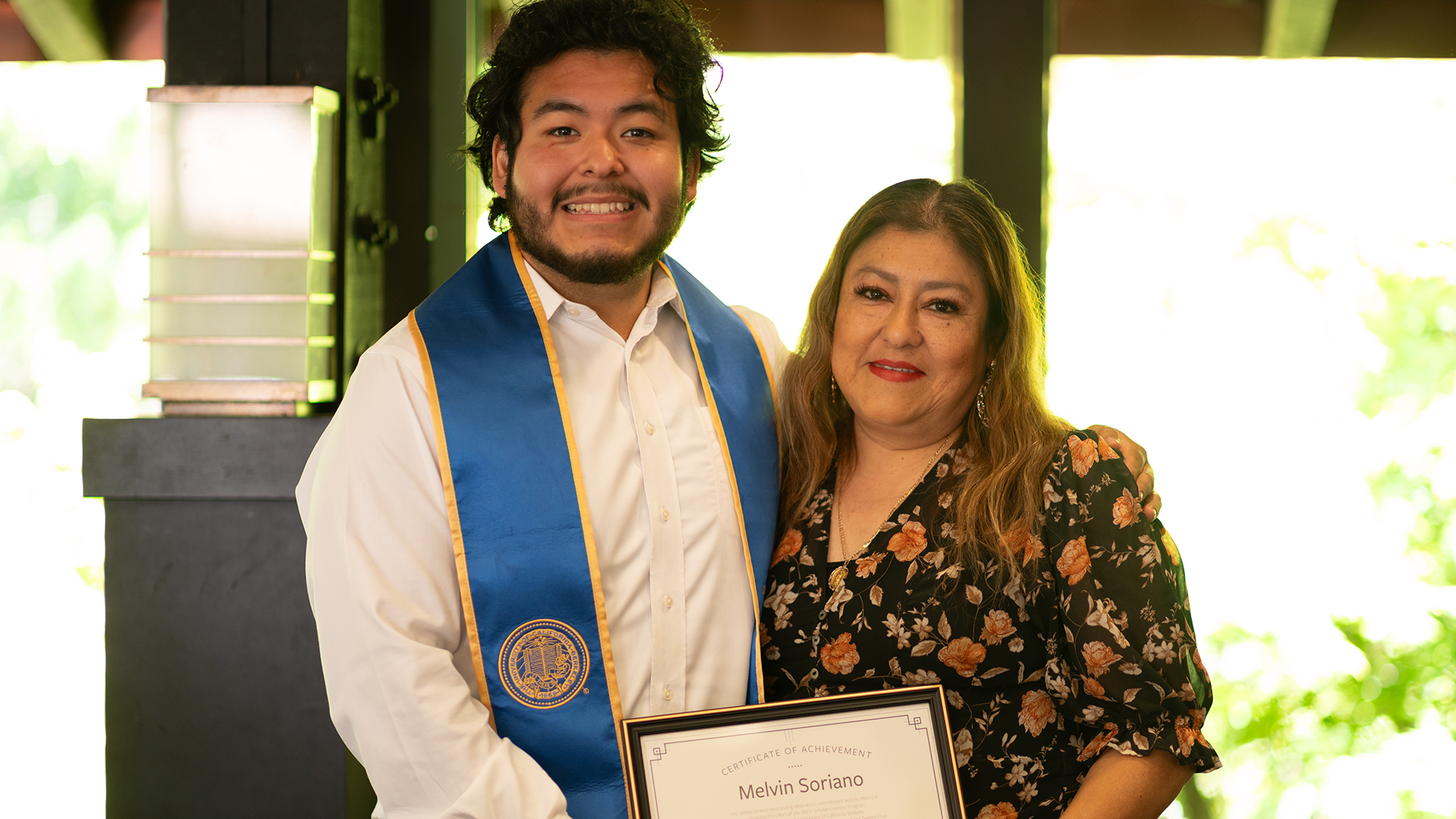 a graduate poses with a loved one holding a framed certificate of achievement