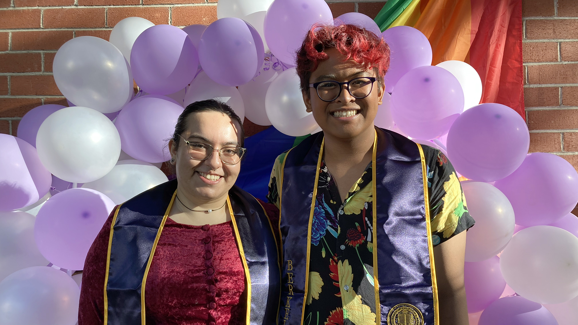 Two graduating students pose together with purple and white balloons and a rainbow flag behind them