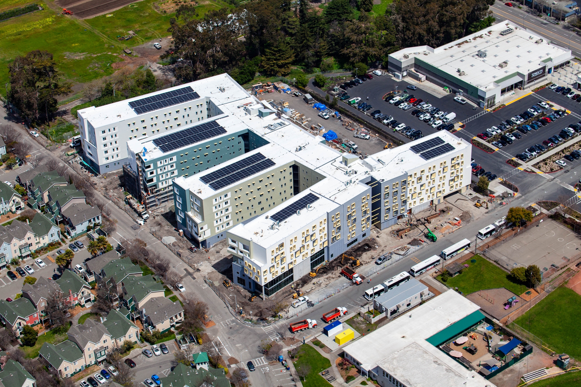 An aerial view of the new UC Berkeley graduate student apartment complex in Albany that is being readied for fall 2024. Work equipment including trucks, caterpillars, tarp, cars and earth being dug up dot the property. Right next to the complex is a Sprouts grocery store and parking lot, other shops and the University Village housing community, for students with families.