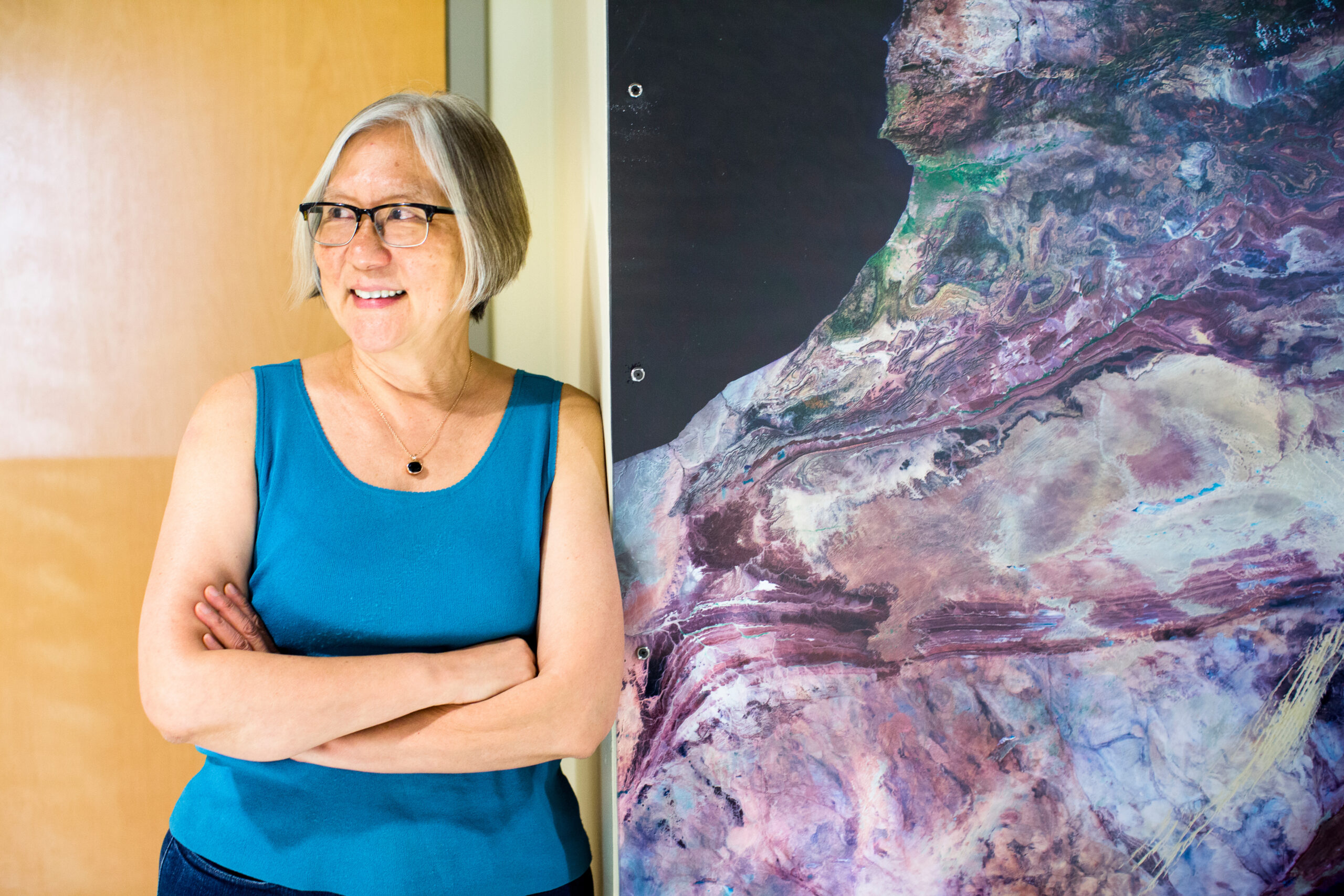 A women wearing a sleeveless blue shirt stands next to a large mural of a purple mineral with her arms crossed