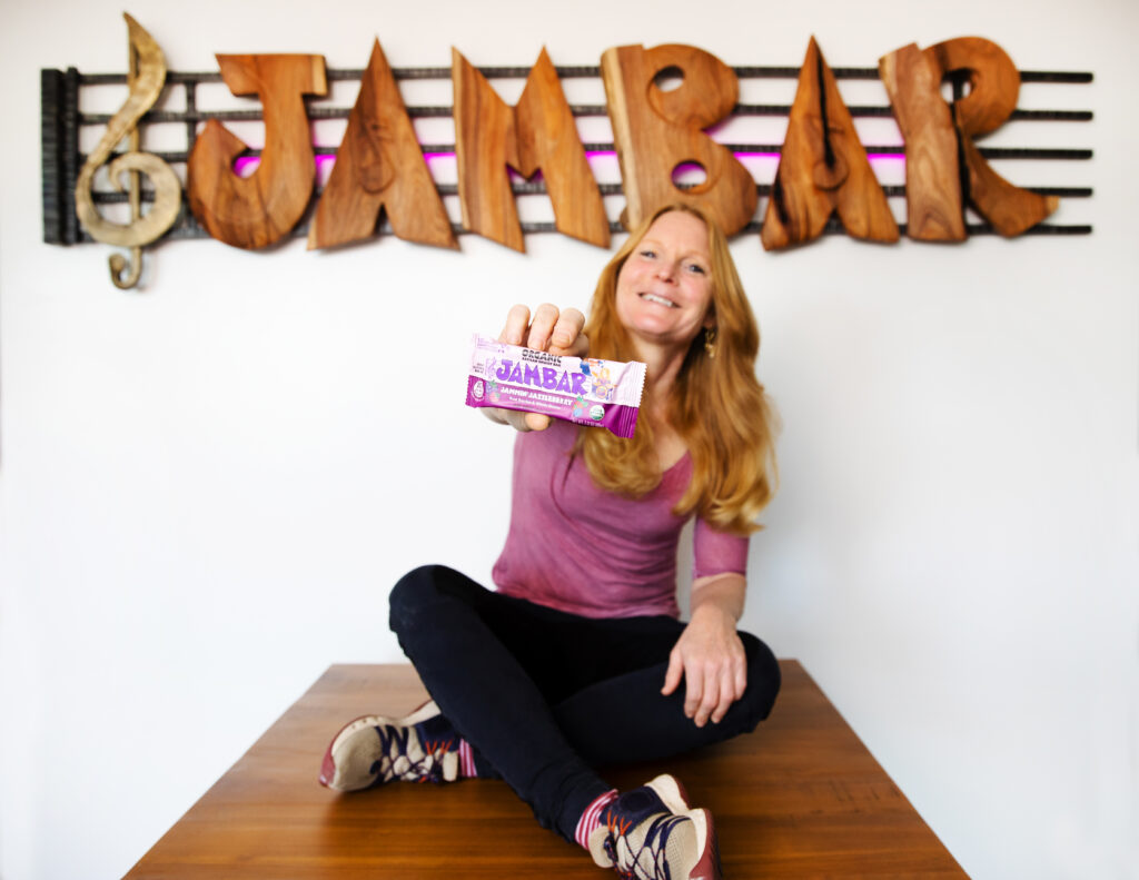 Jennifer Maxwell, present day sitting on a brown table and holding up a JamBar.