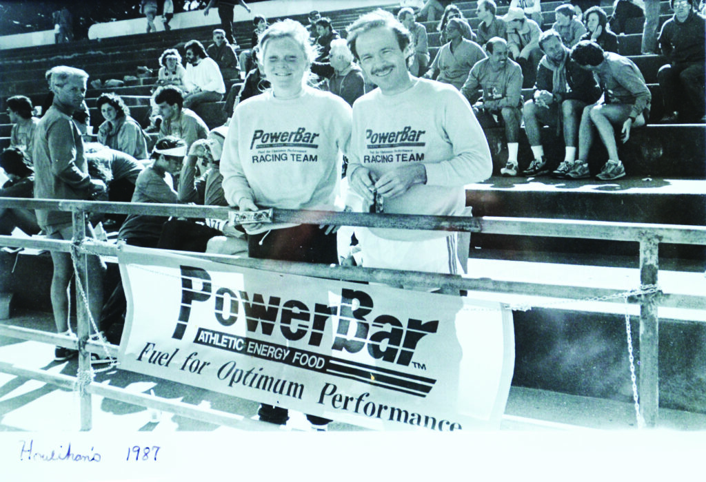 Black and white photo of Jennifer and Brian Maxwell in 1987 with a PowerBar banner hanging on the rail of a track and field event.