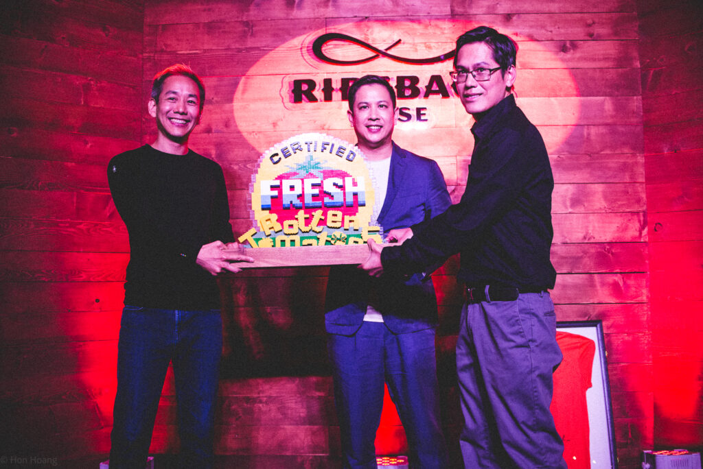 Patrick Lee, Stephan Wang and Senh Duon stand in front of a red lit brick wall as they together hold a trophy that reads certified Fresh Rotten Tomatoes.