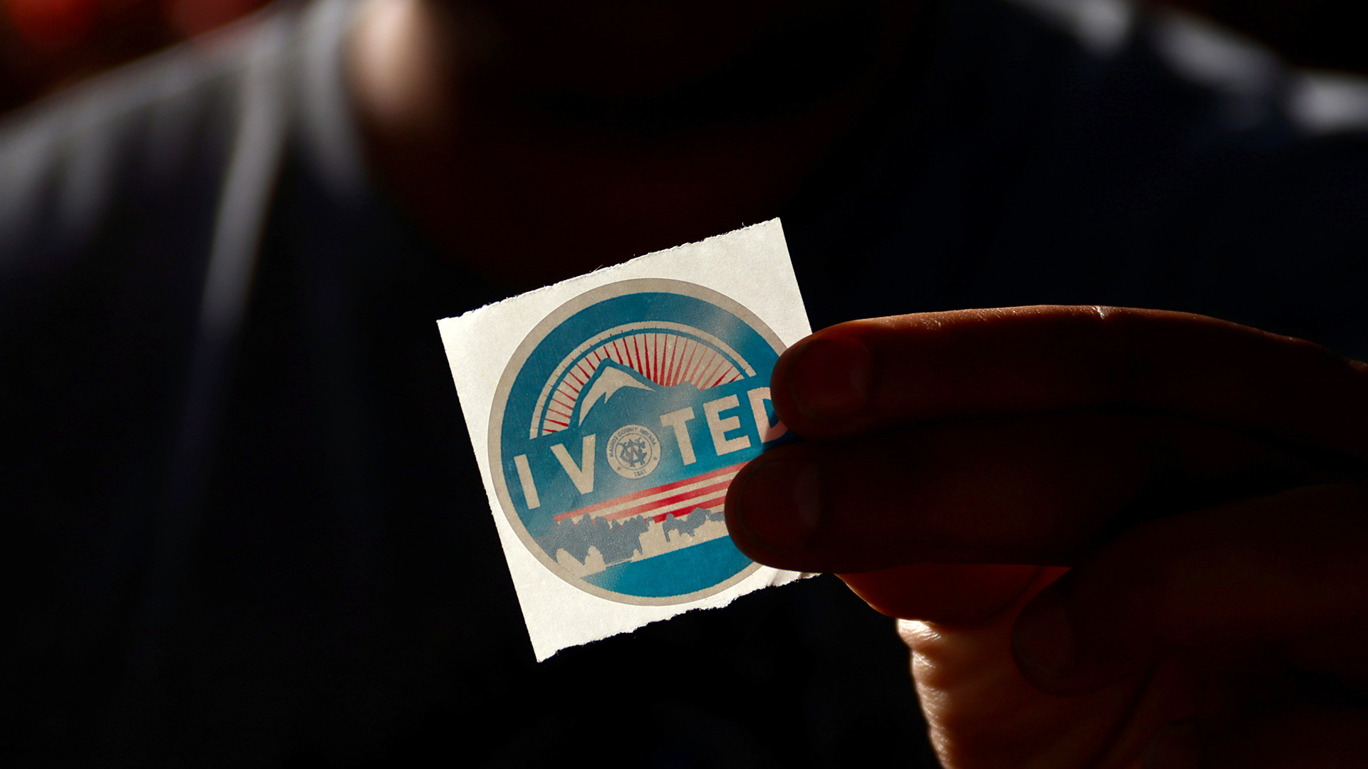 a hand in shadow holds a brightly lit sticker that reads