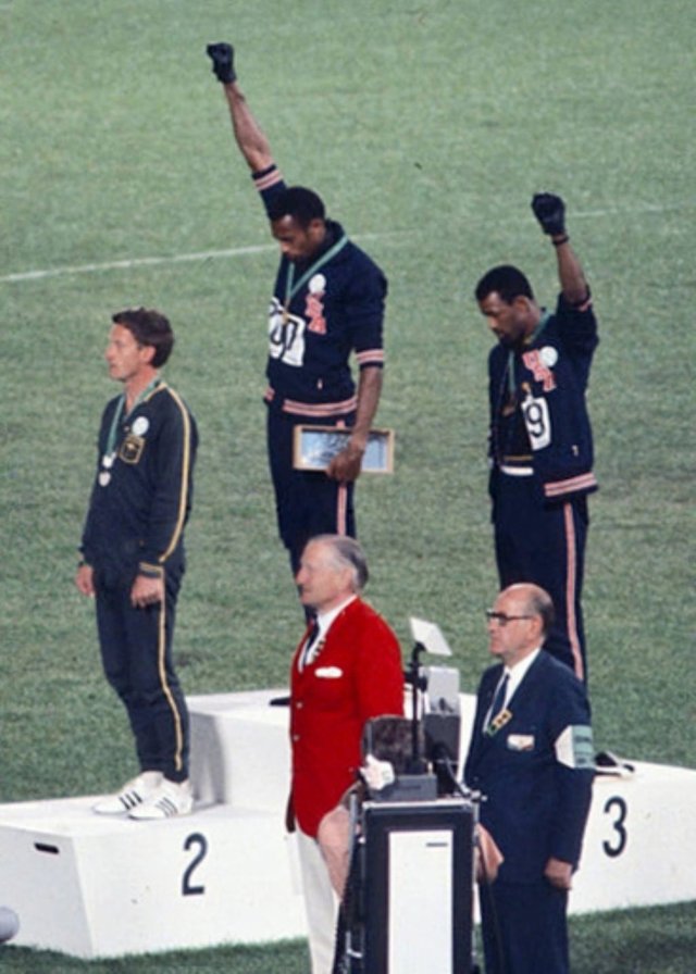 Two Black Olympians Tommie Smith and John Carlos raise a black-gloved fist into the air as they receive medals at an Olympic podium.