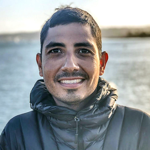 headshot of a person with short dark hair and eyes smiling and wearing a down jacket with a body of water in the background