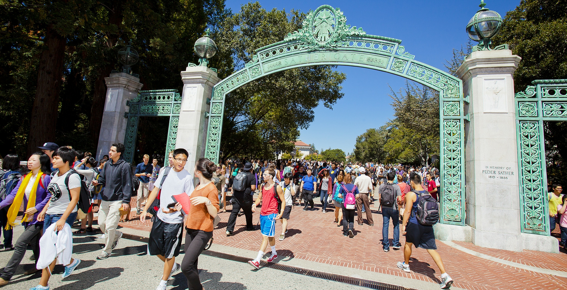 Crowds of students walk under Sather Gate on a warm day, some wearing shorts and sleeveless tops.