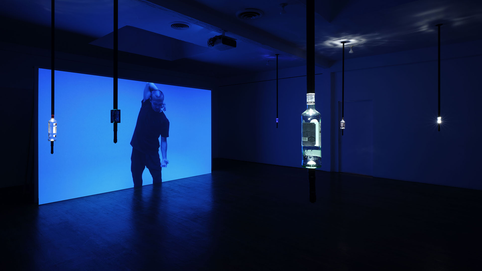 an artist performs a dance in a dark room standing in front of a blue screen with hard alcohol bottles hanging from the ceiling