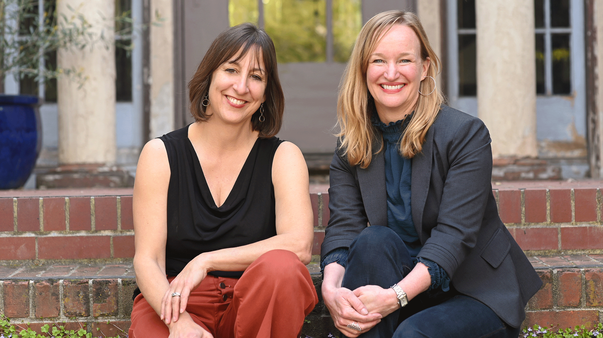 Two women sit side by side on brick steps, smiling