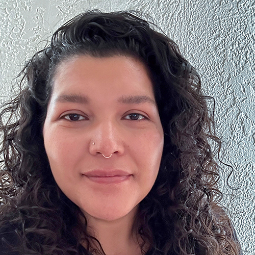 headshot of a person with long curly hair and a nose piercing smiling slightly