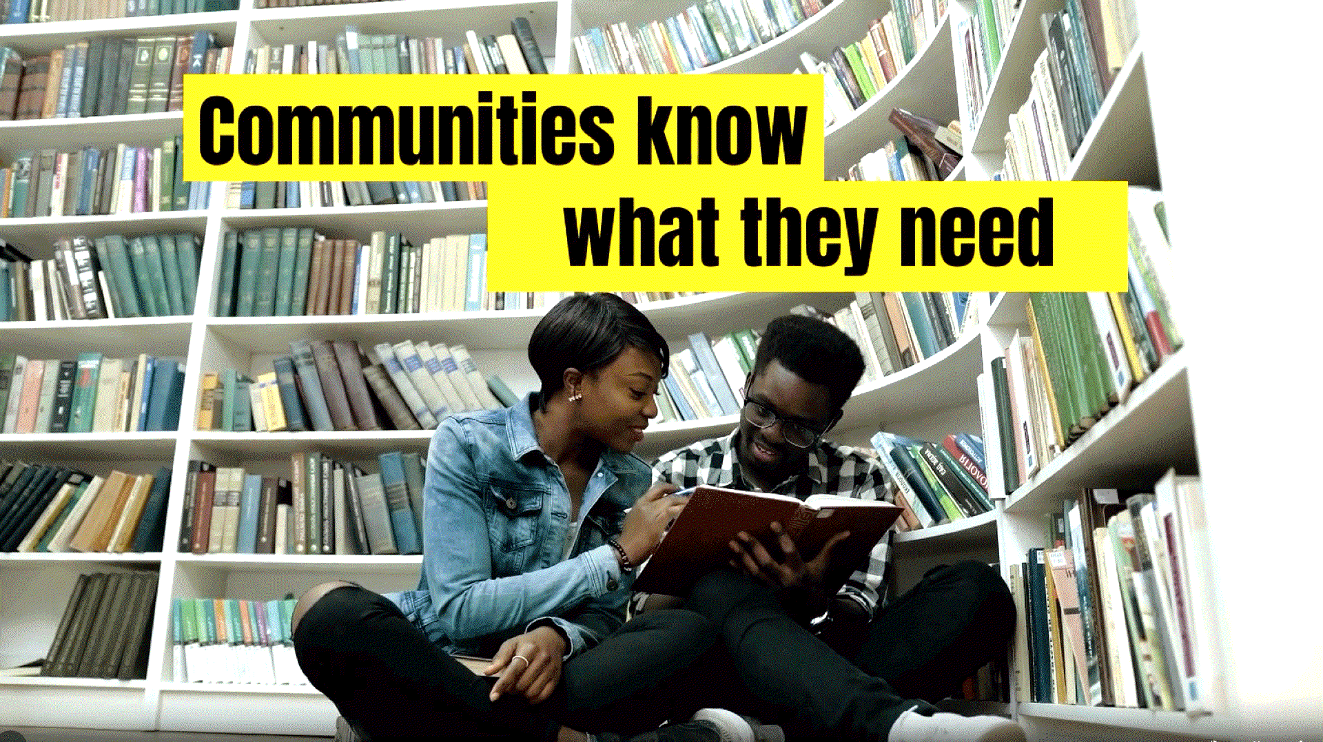 Two people sit cross-legged on the floor of a library, smiling as they read a book