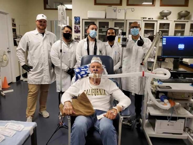 an older man in a California T-shirt sitting in a chair with a tube connected to his mouth, with five people in white coats and masks behind him