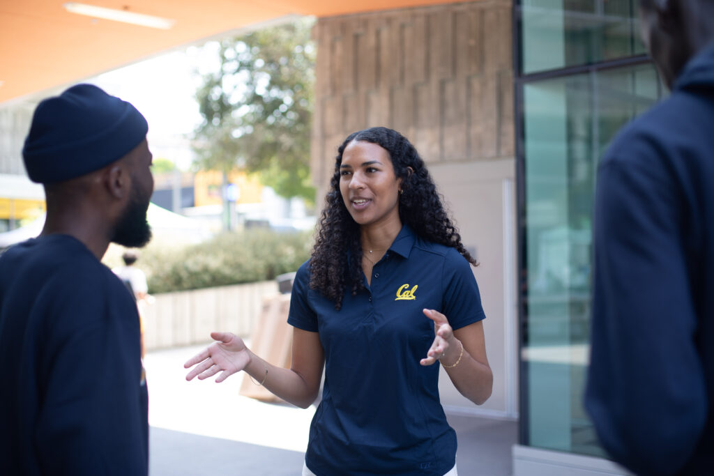 Sydney Roberts, the new ASUC president, is outside the ASUC Student Union meeting two students with her hands outstretched as she talks. She is wearing a dark blue polo shirt with the word Cal on it.