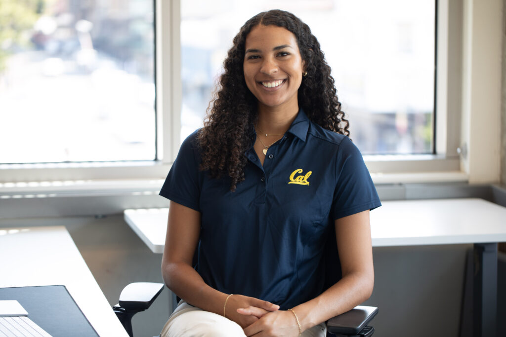 Sydney Roberts, the new ASUC president, sits at a desk in the ASUC student union and faces the camera. She is smiling, with long curly hair and a dark blue polo shirt with Cal written on it.