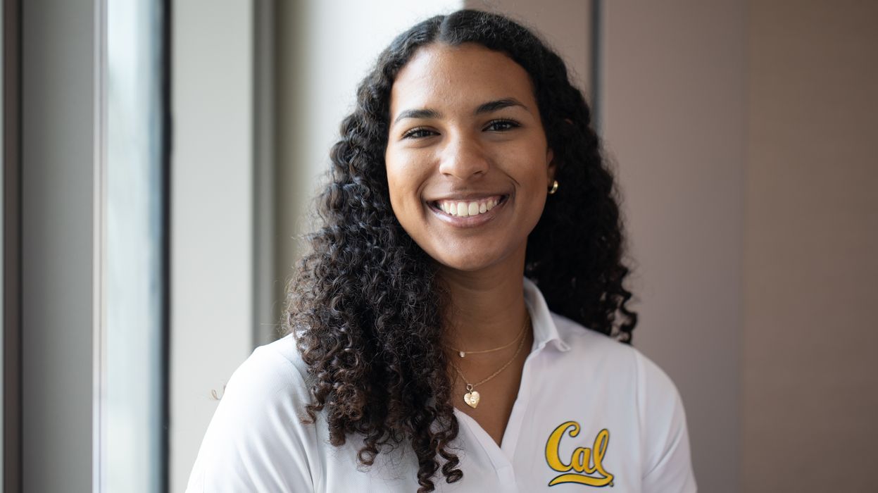Sydney Roberts, the new ASUC president, poses for a casual portrait. She has a wide smile, a white polo shirt with Cal written on it, gold jewelry and long curly hair.