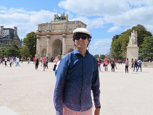 Saagar, wearing casual clothing and a beret, stands in a public space outside with the Louvre in Paris in the background