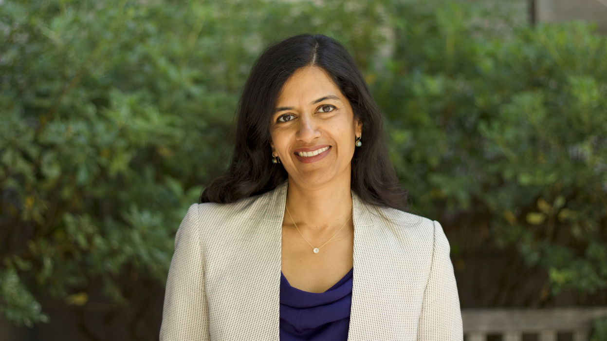 A headshot of Manisha Shah, smiling, outside in front of bushes