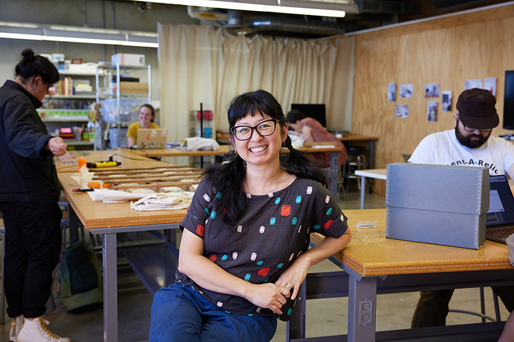 A Filipinx American woman with long dark hair and glasses wearing a colorful shirt smiles while she sits at a table in an art classroom while students work on projects in the background