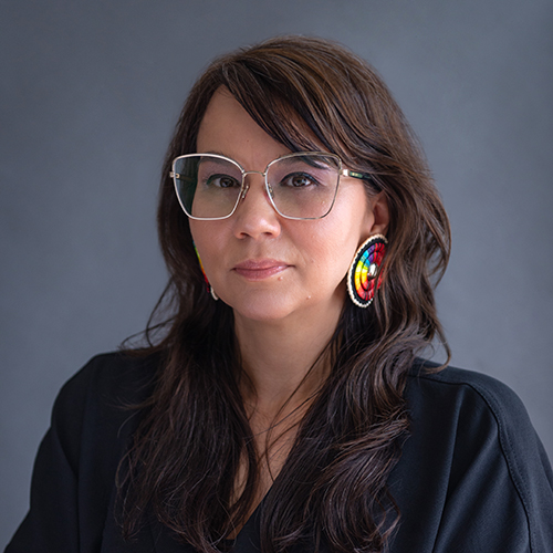 headshot of a person with long wavy hair wearing glasses and colorful earrings