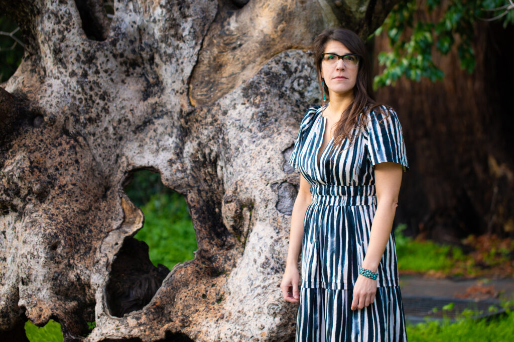 Tedde Simon, the campus's new tribal liaison, stands in front of the iconic old California Buckeye tree in Faculty Glade wearing a vertically-striped dress and turquoise jewelry.