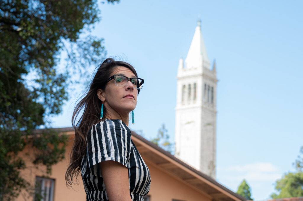 Tedde Simon, the campus's new tribal liaison, looks sideways at the camera with the Campanile in the distance. She's wearing a vertically-striped dress and long turquoise earrings.