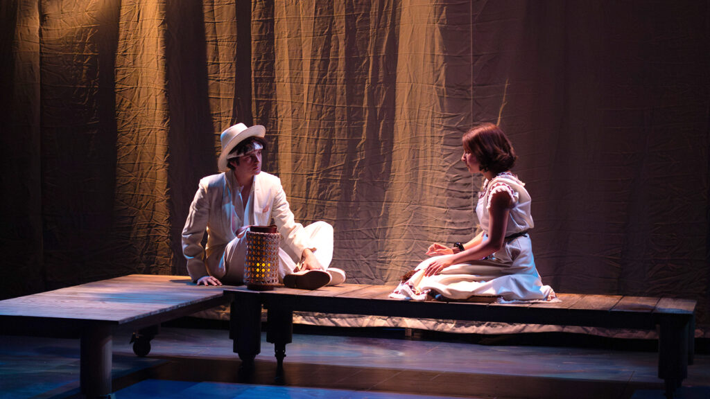 Two actors sit closely facing each other and talk on a wooden platform representing a dock 