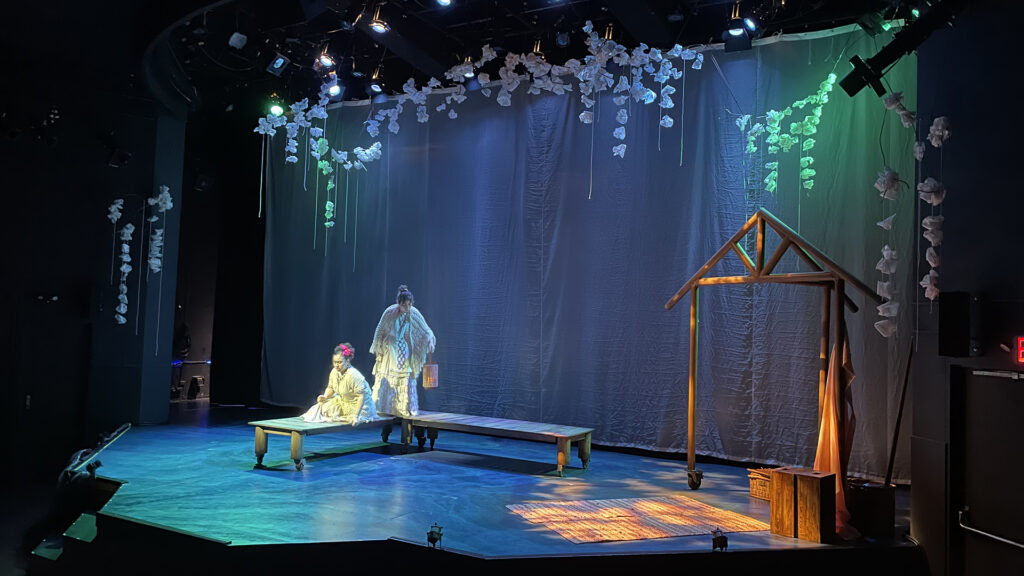 A theater stage lit with purple and green lights. Two actors are on a wooden platform that represents a dock in the water.