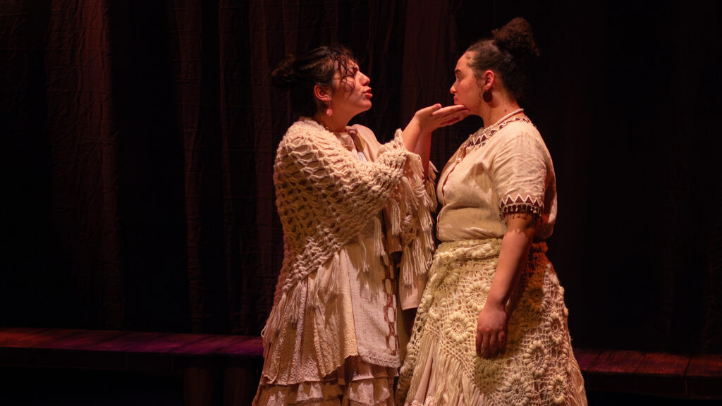 Two actors playing mother and daughter stand on stage, both wearing off-white, woven-looking dresses. The mother, wearing a shawl, kind of cups the chin of her daughter, in an attempt for her to look at her and listen to what she's saying.