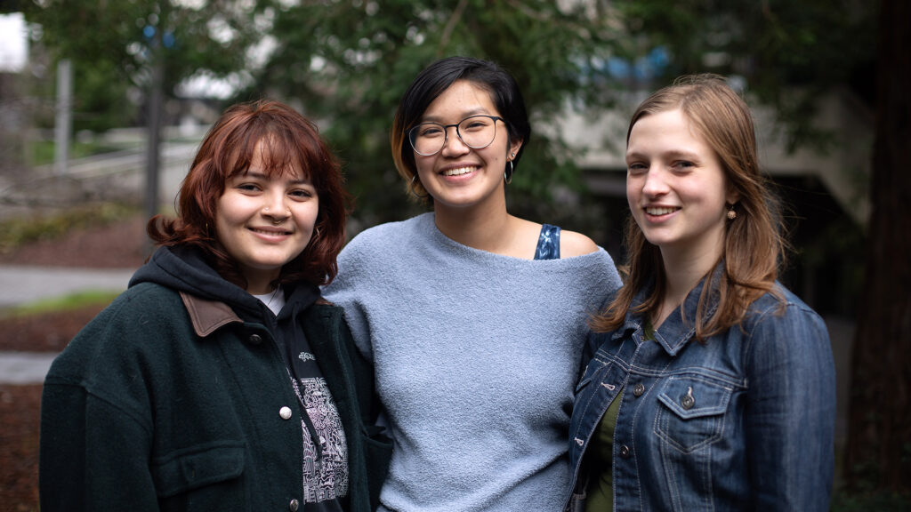 Students Lee, Amy and Mirin stand close to one another side-by-side and smile for a portrait