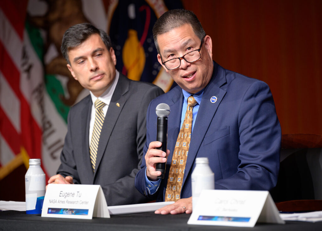 two men at table, one speaking into a microphone