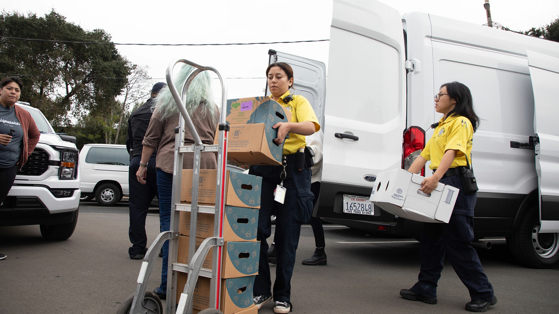 Volunteers wearing yellow shirts and black pants unload boxes from the back of a white van and stack them onto a dolly.