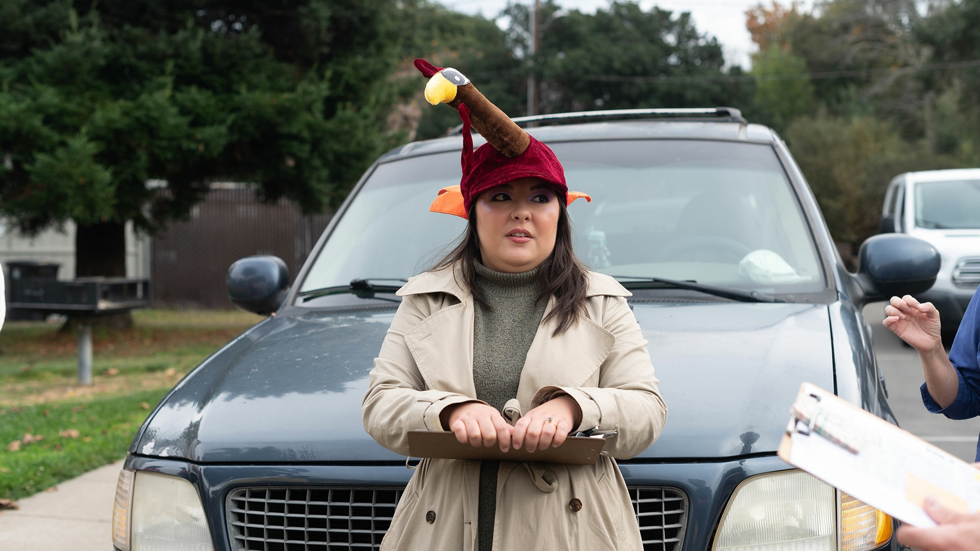 A volunteer wearing a turkey hat holds a clipboard and leans against a car.
